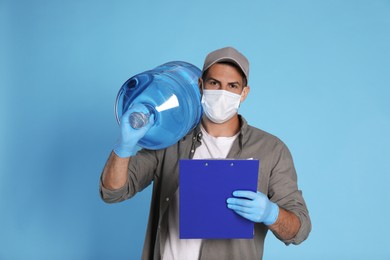 Photo of Courier in medical mask with bottle for water cooler and clipboard on light blue background. Delivery during coronavirus quarantine