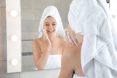 Photo of Young woman washing face with soap near mirror in bathroom