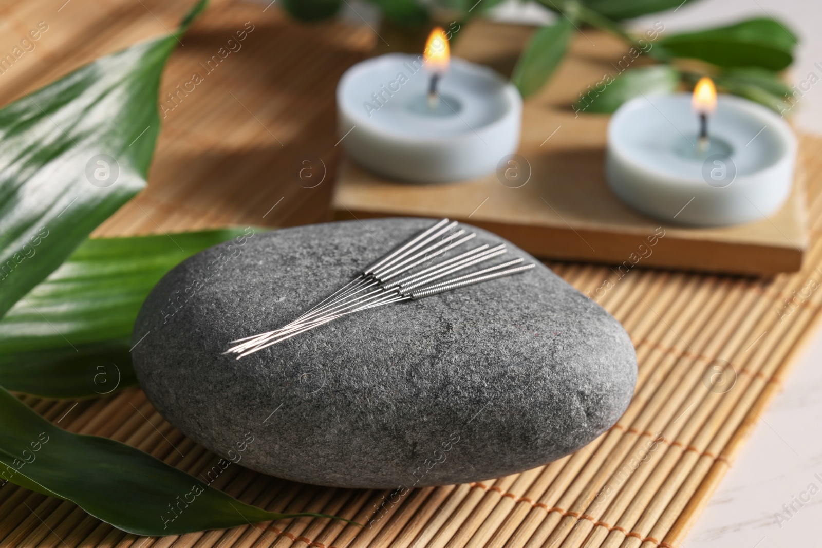 Photo of Stone with acupuncture needles and burning candles on table, closeup