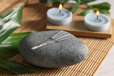 Photo of Stone with acupuncture needles and burning candles on table, closeup