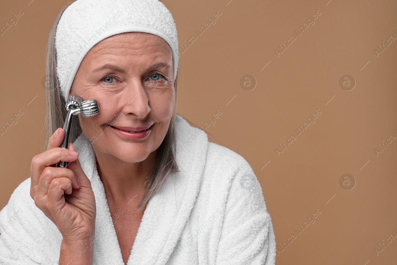 Photo of Woman massaging her face with metal roller on brown background. Space for text