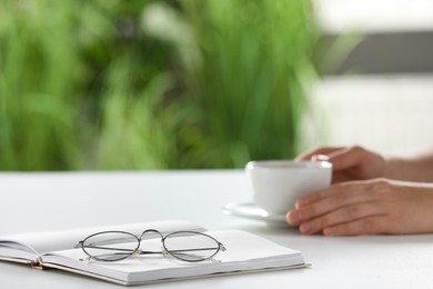 Photo of Woman holding cup of coffee at white table indoors, focus on eyeglasses and notebook