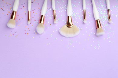 Photo of Different makeup brushes and shiny confetti on violet background, flat lay. Space for text