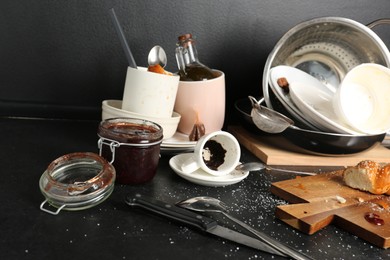 Many dirty utensils, dishware and food leftovers on black countertop. Mess in kitchen