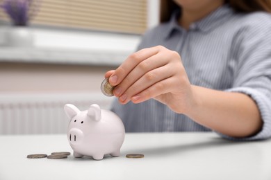 Woman putting coin into ceramic piggy bank at white wooden table indoors, closeup. Financial savings