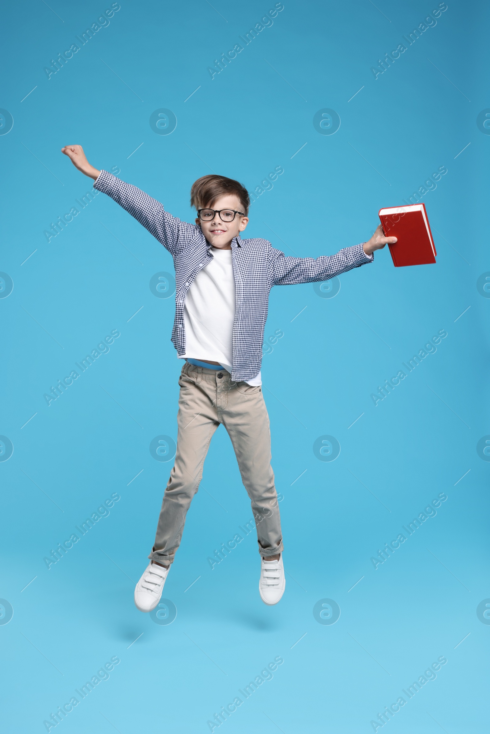 Photo of Cute schoolboy in glasses holding book and jumping on light blue background