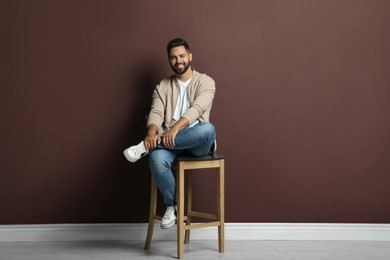 Photo of Handsome young man sitting on stool near brown wall