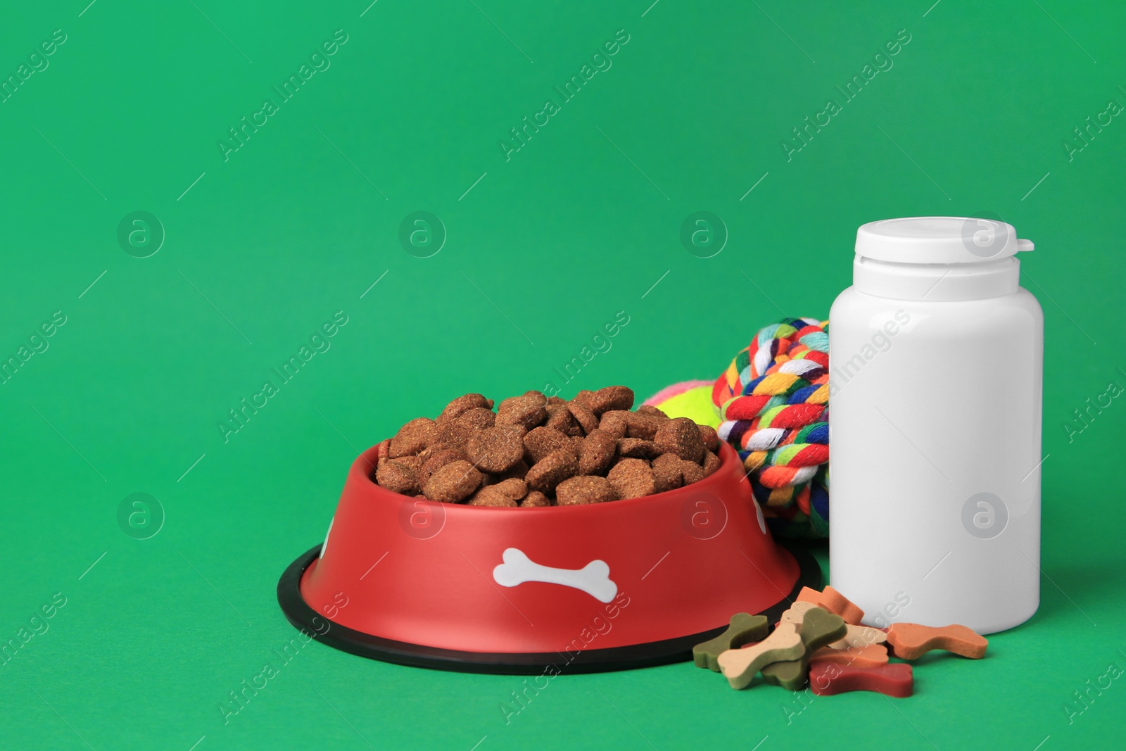 Photo of Bowl with dry pet food, bottle of vitamins and toys on green background. Space for text