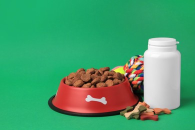 Photo of Bowl with dry pet food, bottle of vitamins and toys on green background. Space for text