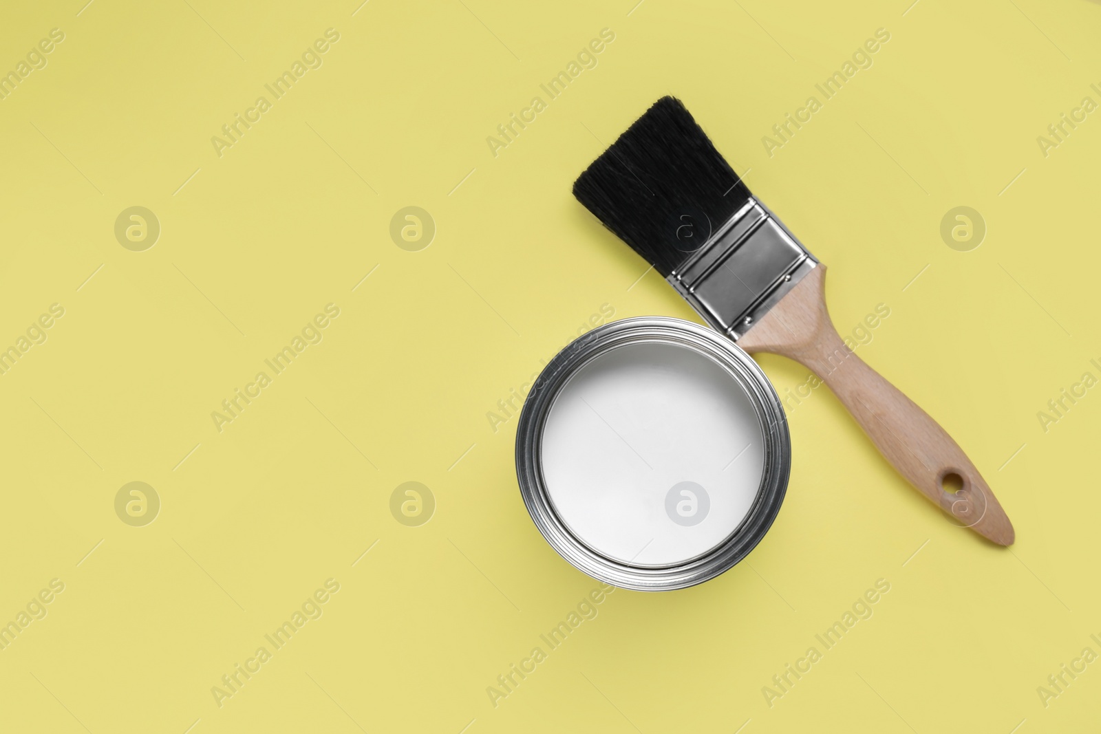 Photo of Can with white paint and brush on yellow background, flat lay. Space for text