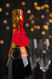 Heart shaped greeting card with phrase I Love You tied to bottle of champagne against blurred lights, closeup