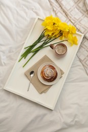 Bouquet of beautiful daffodils, bun and coffee on bed, top view