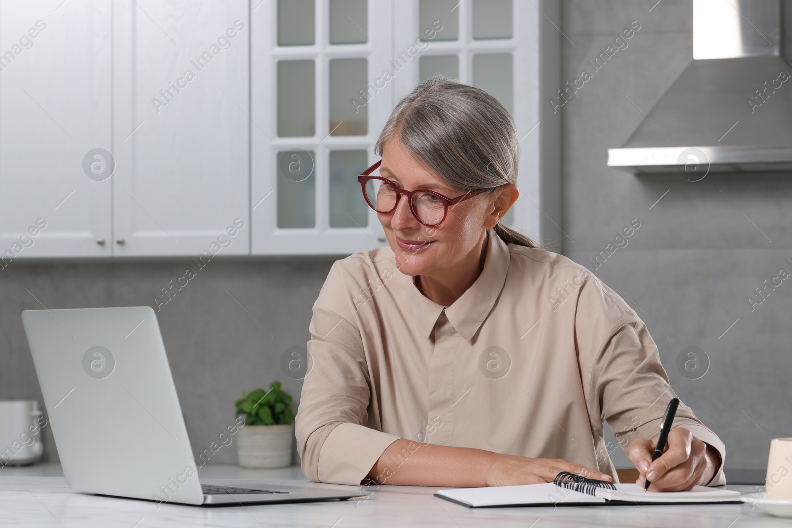 Photo of Beautiful senior woman taking notes while using laptop at white marble table in kitchen