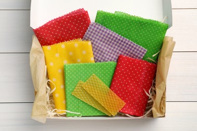 Photo of Colorful beeswax food wraps in box on white wooden table, top view