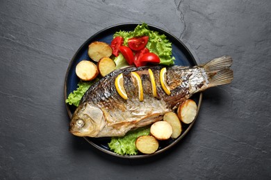 Photo of Tasty homemade roasted crucian carp with garnish on black table, top view. River fish