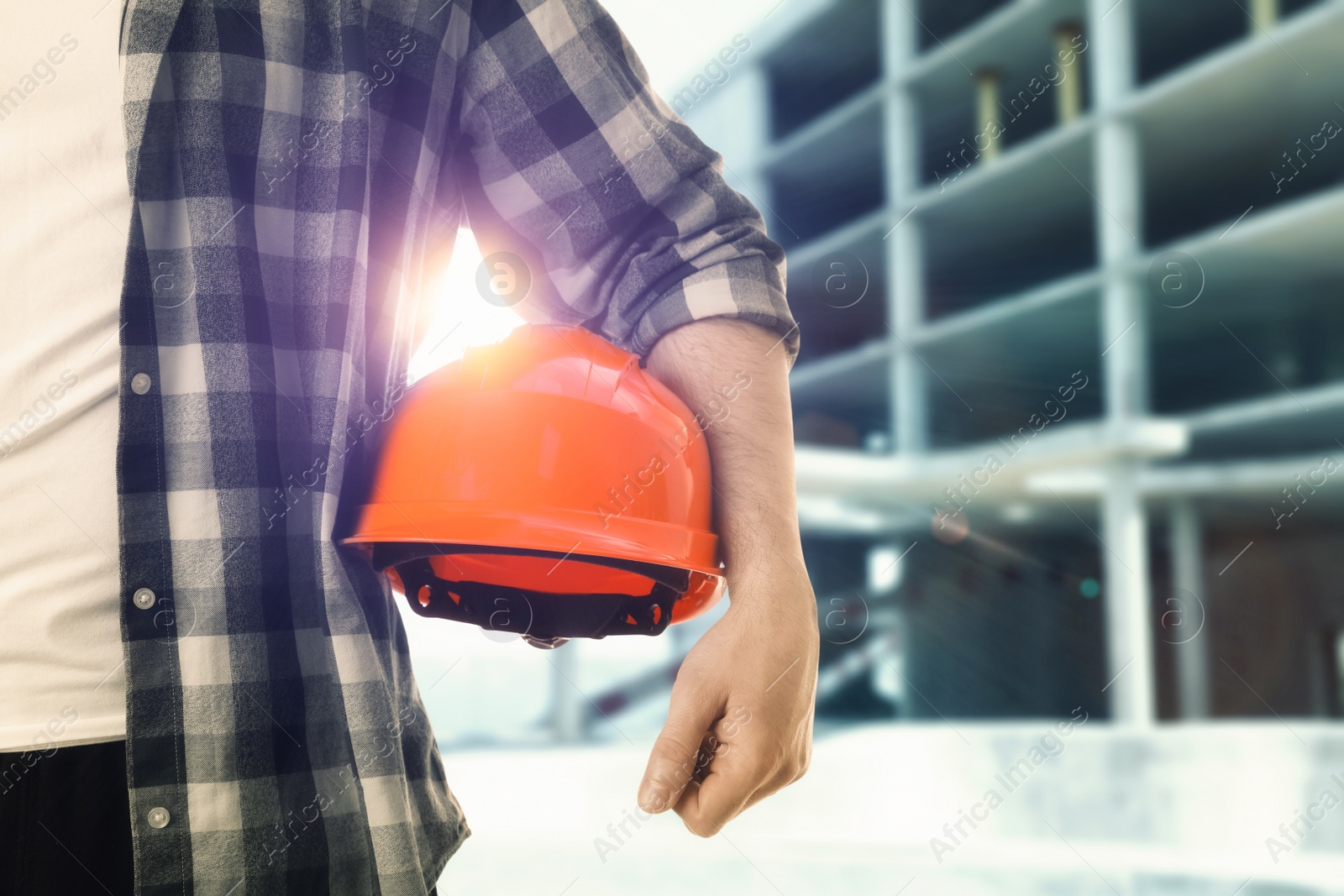 Image of Man holding orange hard hat at construction site with unfinished building, closeup. Space for text 