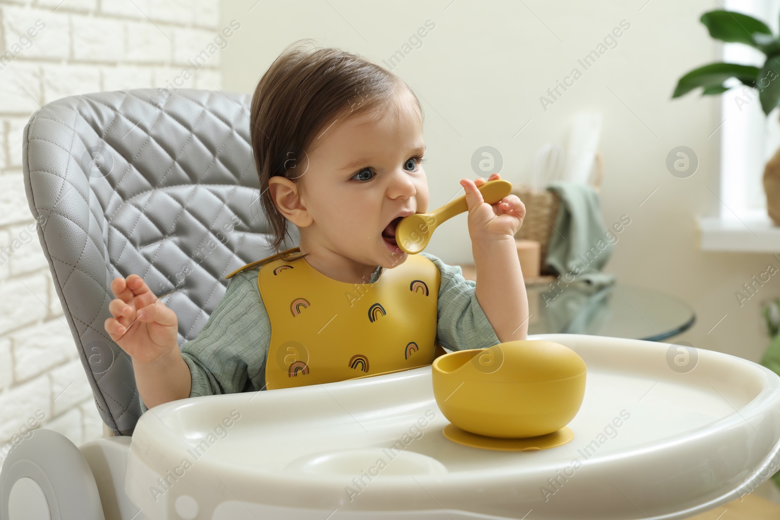 Photo of Cute little baby nibbling spoon in high chair indoors.