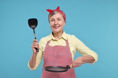 Happy housewife with turner and frying pan on light blue background