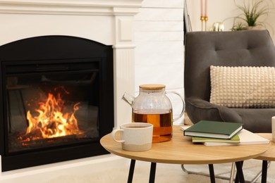 Teapot with cup of hot drink and books on wooden table near decorative fireplace in room. Interior design