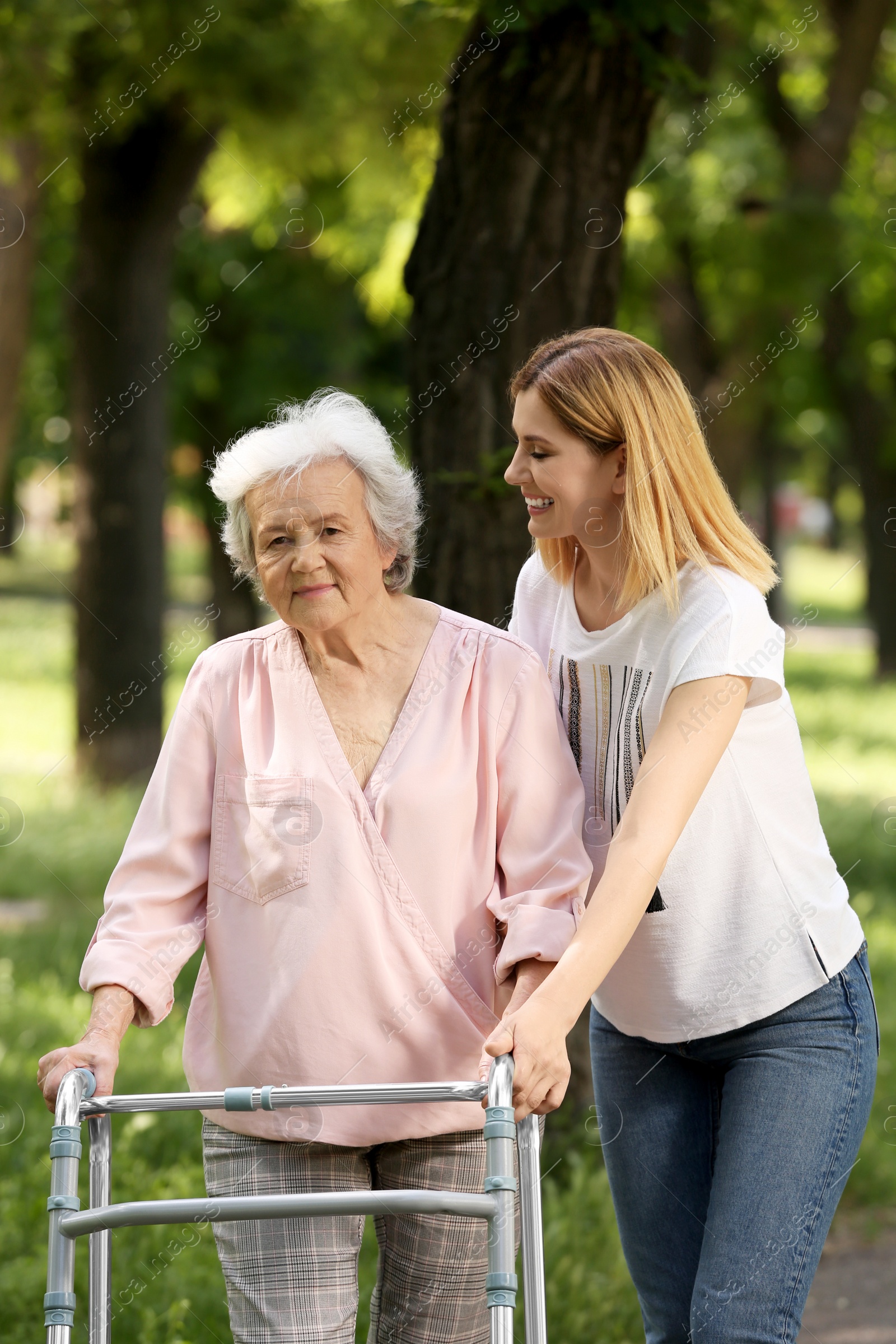 Photo of Caretaker helping elderly woman with walking frame outdoors