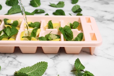 Photo of Pouring water into ice cube tray with mint and lemon on marble table