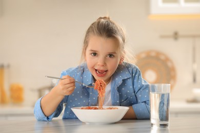 Photo of Cute little girl eating tasty pasta at table in kitchen