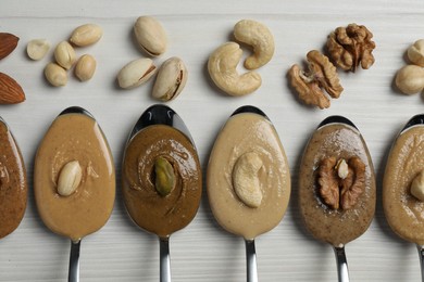 Tasty nut butters in spoons and raw nuts on white wooden table, flat lay