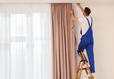 Photo of Worker in uniform hanging window curtain indoors, back view