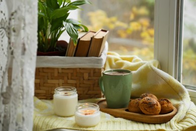 Photo of Burning aromatic candles, cup with hot drink and cookies on windowsill indoors. Autumn coziness