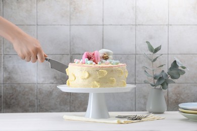 Woman cutting delicious cake decorated with macarons and marshmallows at white wooden table, closeup