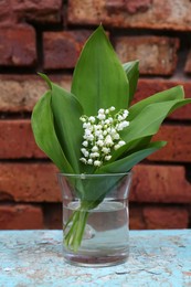 Photo of Beautiful lily of the valley flowers in glass vase on turquoise wooden table near brick wall