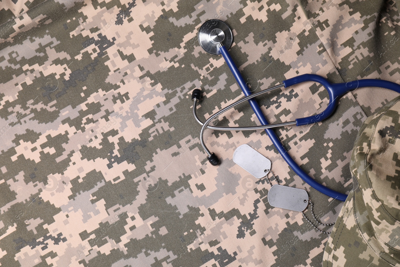 Photo of Stethoscope, military ID tags and cap on camouflage fabric, flat lay. Space for text