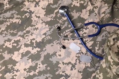 Photo of Stethoscope, military ID tags and cap on camouflage fabric, flat lay. Space for text