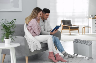 Photo of Happy couple sitting on sofa near electric heater at home