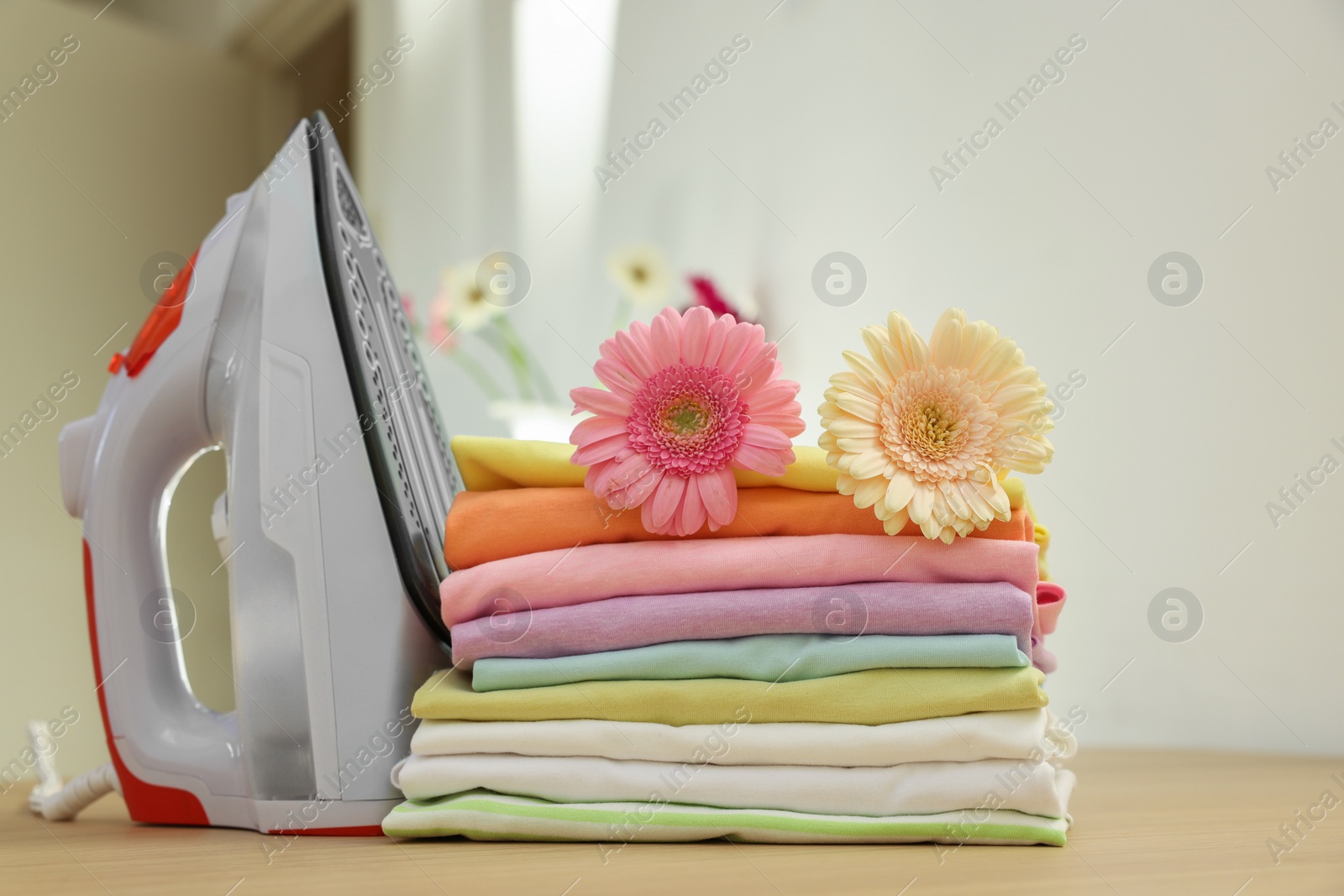 Photo of Stack of clean clothes, modern iron and flowers on wooden table