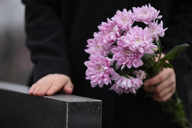 Photo of Woman holding chrysanthemum flowers near black granite tombstone outdoors, closeup. Funeral ceremony