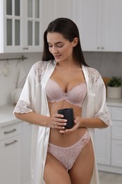 Young woman with cup wearing elegant underwear and robe in kitchen