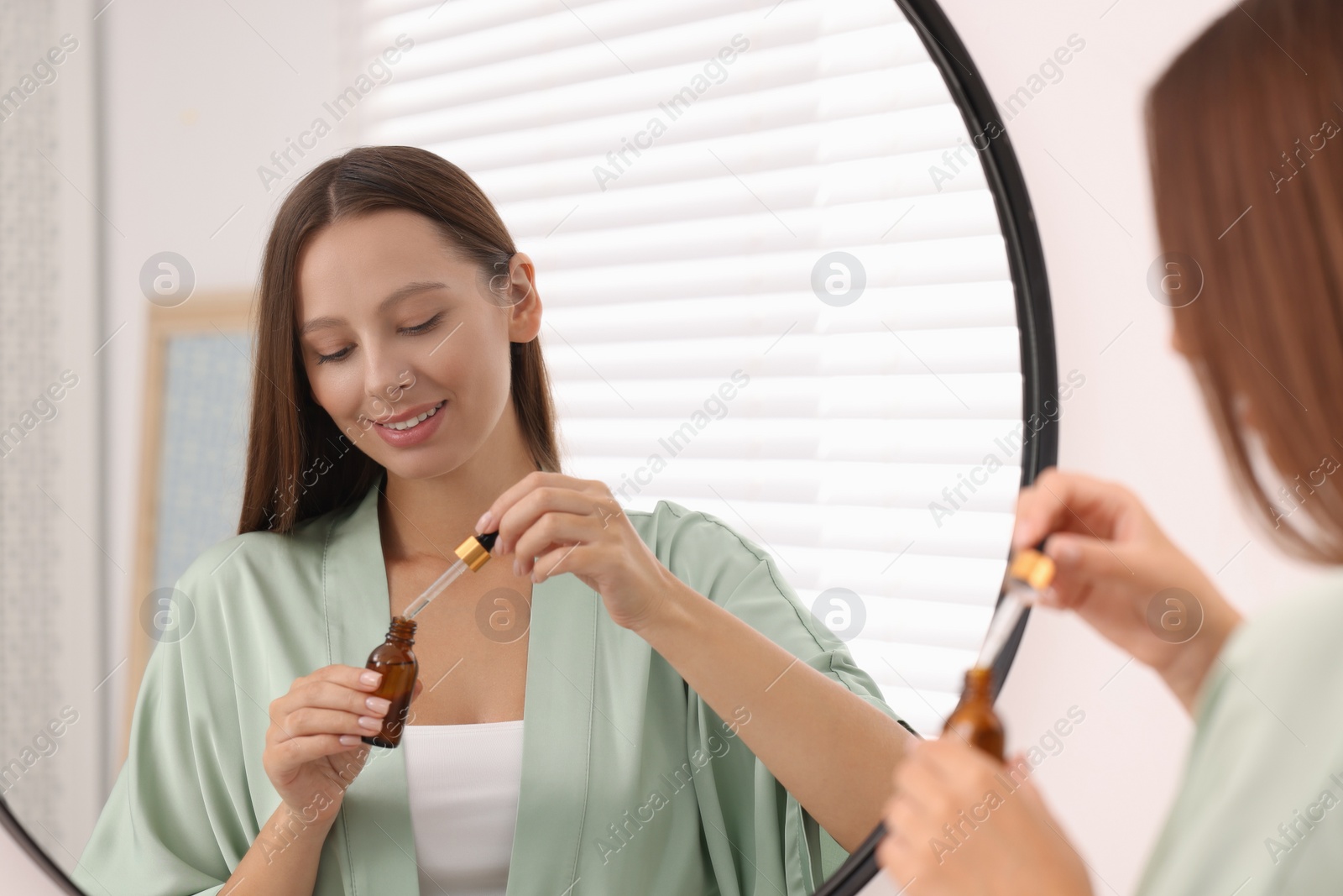 Photo of Beautiful woman holding bottle of serum and dropper near mirror indoors