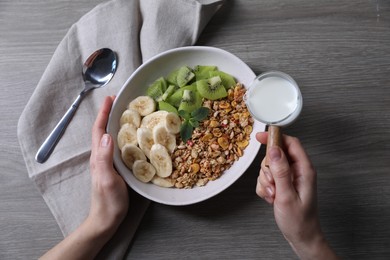 Woman pouring milk into bowl of tasty granola with banana and kiwi at grey wooden table, top view