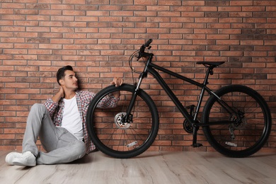 Handsome young man with modern bicycle near brick wall indoors