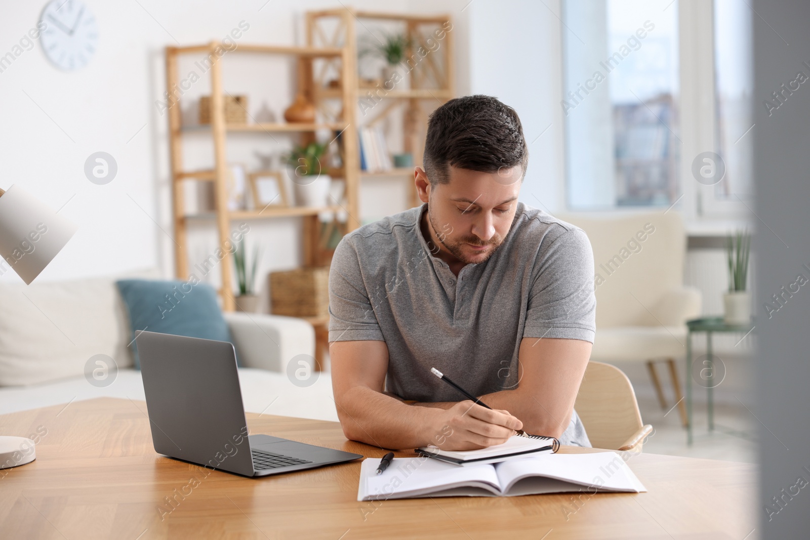 Photo of Man writing notes while working on laptop at wooden desk in room