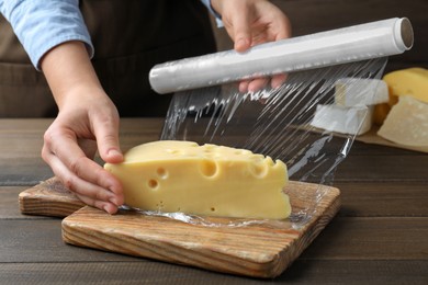 Photo of Woman putting plastic food wrap over block of cheese at wooden table, closeup
