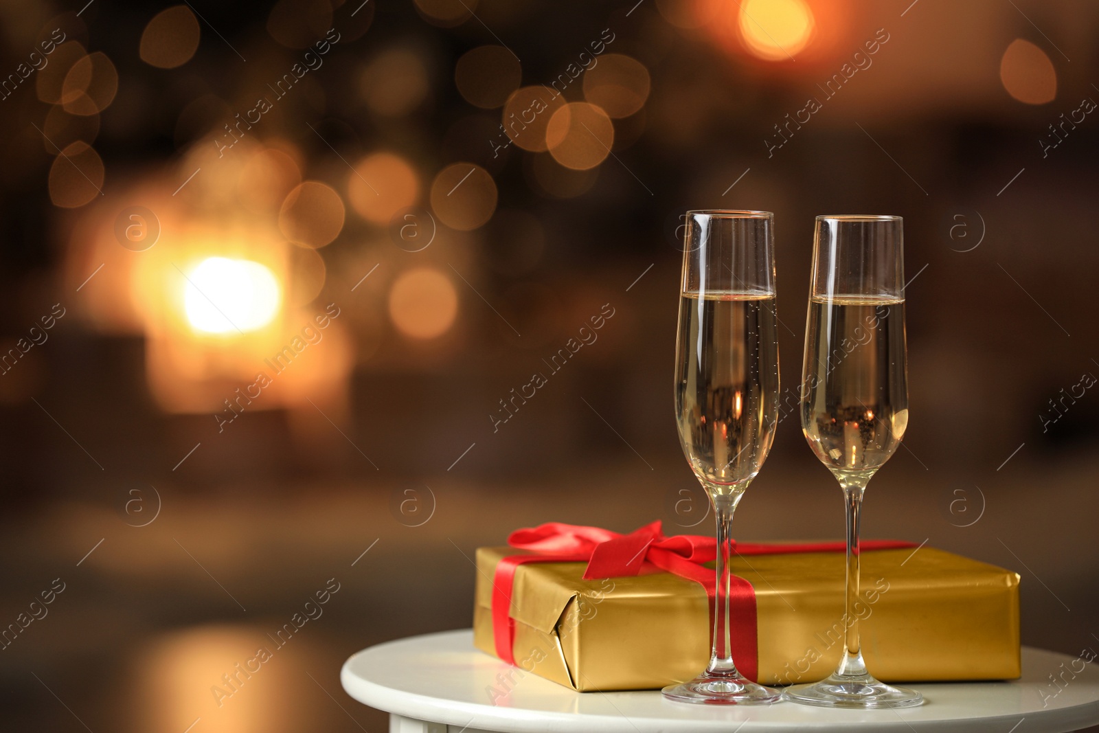 Image of Glasses with champagne and Christmas gift on table in room, bokeh effect. Space for text