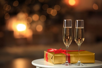 Image of Glasses with champagne and Christmas gift on table in room, bokeh effect. Space for text