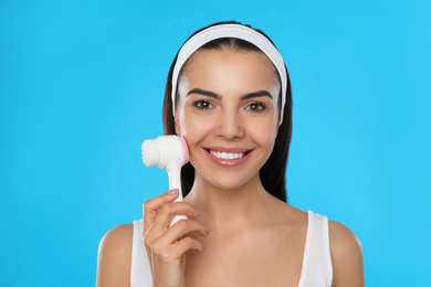 Photo of Young woman using facial cleansing brush on light blue background. Washing accessory