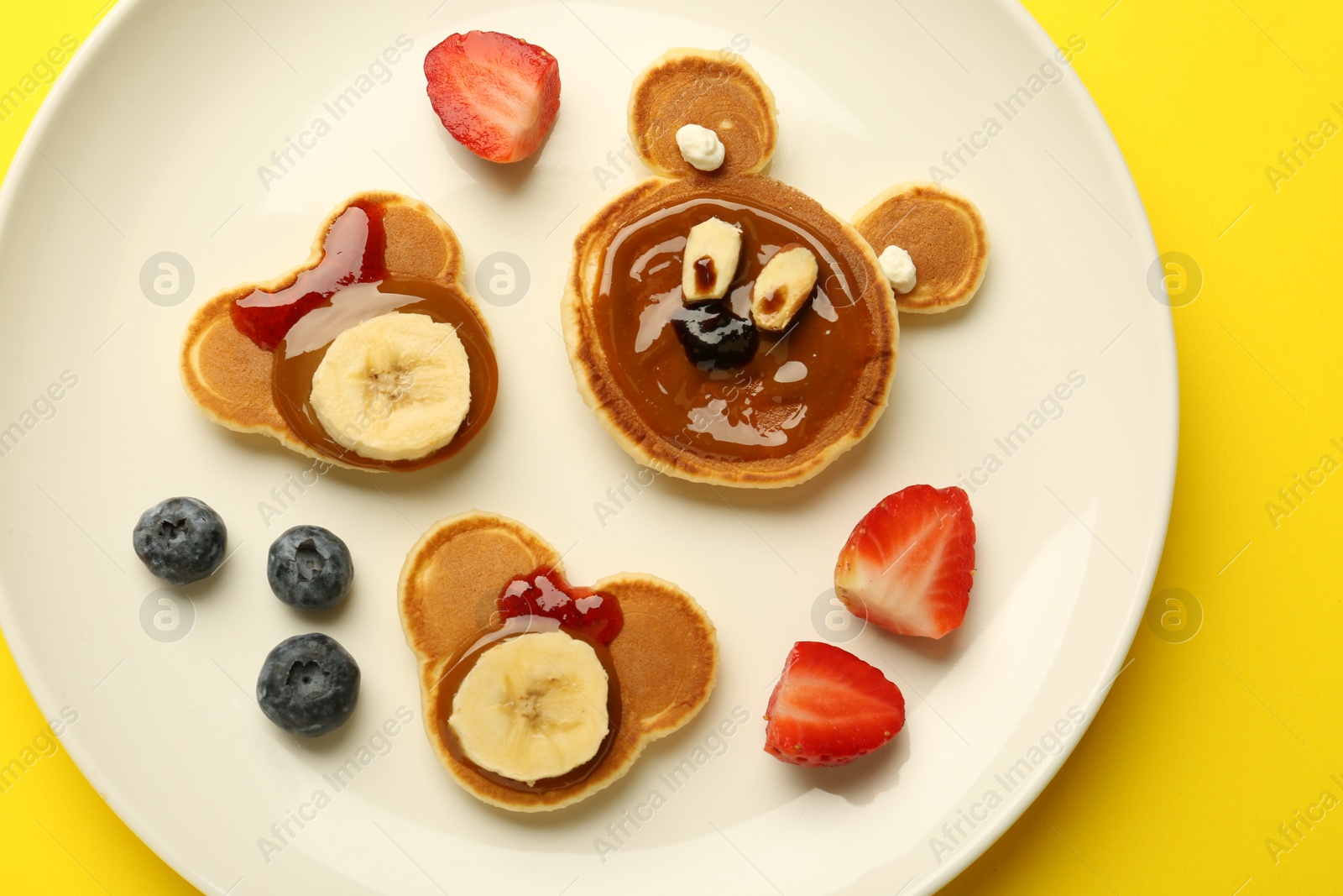 Photo of Creative serving for kids. Plate with cute bears made of pancakes, berries, banana and chocolate paste on yellow background, top view