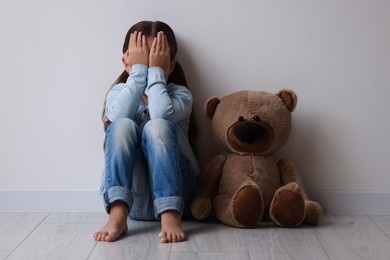 Photo of Child abuse. Upset little girl with teddy bear sitting on floor near light wall indoors