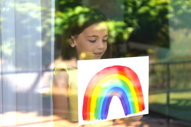 Photo of Little girl with picture of rainbow near window, view from outdoors. Stay at home concept