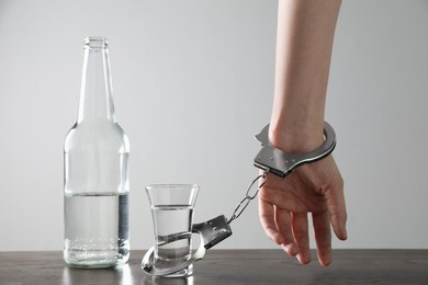 Photo of Woman in handcuffs with glass of vodka at wooden table against white background, closeup. Alcohol addiction