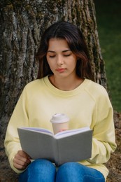 Young woman with cup of coffee reading book near tree in park